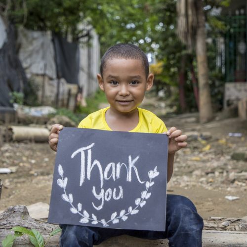 Een jongen in een geel shirt houdt een bord vast met de tekst Thank you.